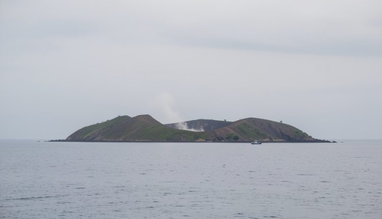 A low volcanic island in the Sunda Strait seen from the water with a thin wisp of steam from the crater and a distant patrol boat on a calm grey sea