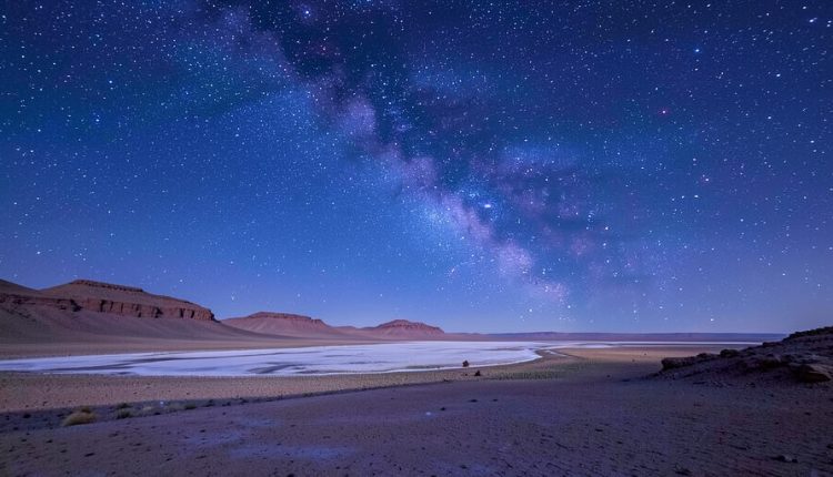 The Milky Way arching over a barren high-altitude desert landscape with distant hills and a salt flat under a star-filled sky