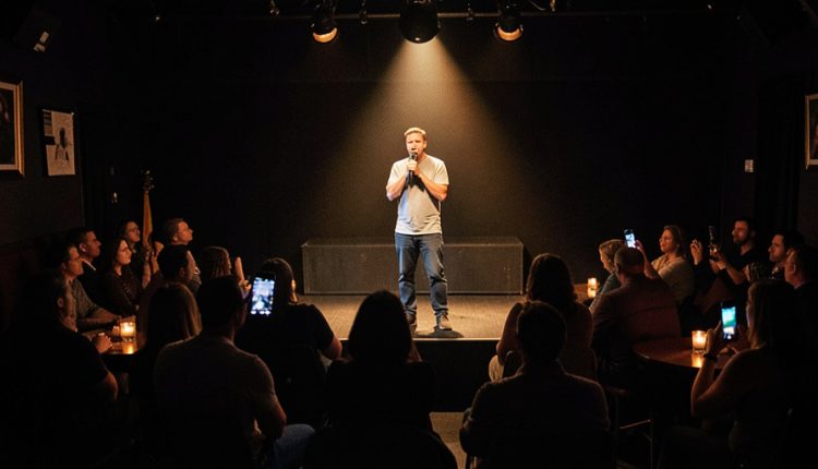 A stand-up comedian in a spotlight on a small club stage while several audience members hold up glowing phones to record