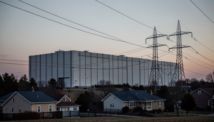 Large windowless data center building behind suburban houses at dusk with high-voltage transmission lines