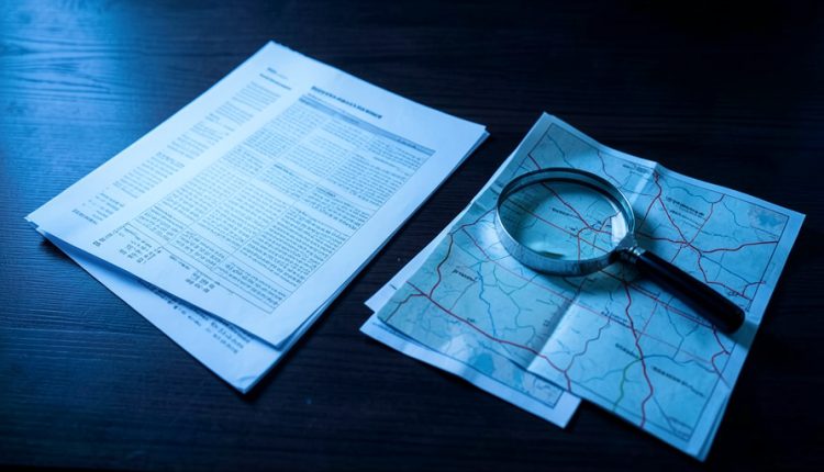Printed spreadsheets and a road map with a magnifying lens on a dark desk under cool blue light