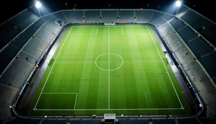 Overhead view of a floodlit football pitch at night showing the center circle and pitch markings, with empty stands around it