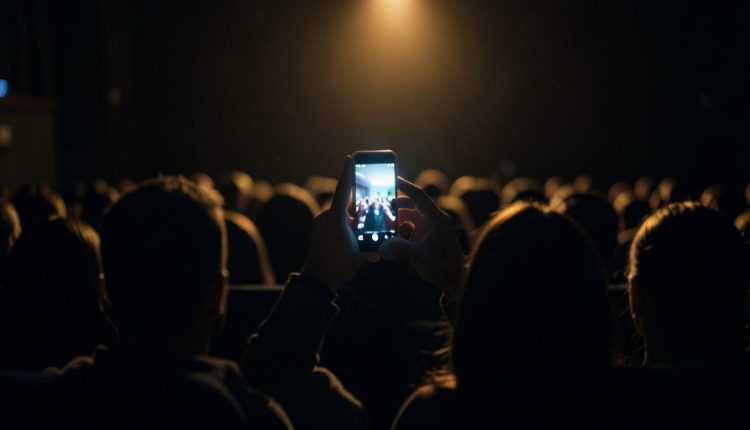 A single glowing smartphone raised to film in a dark theater, seen from behind over the silhouetted heads of an audience