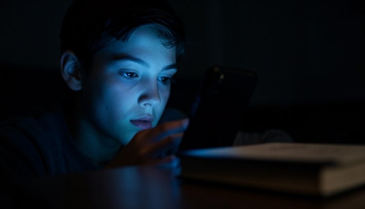 Teenager's face lit by the blue glow of a smartphone in a dark room with a closed book set aside