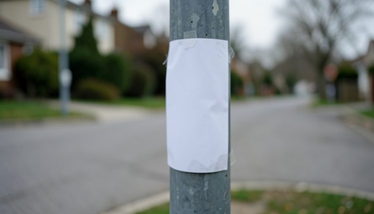 A blank white paper flyer taped to a lamppost on a quiet residential street under overcast daylight