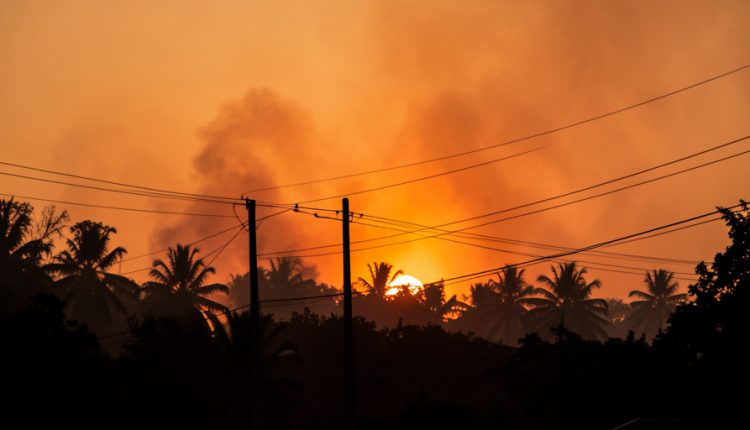 Power lines and utility poles silhouetted against a hazy orange smoke-filled dusk sky over a tropical forest