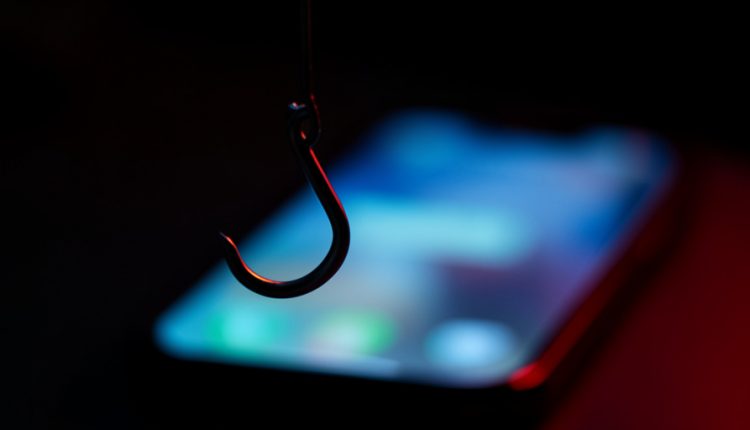 A fishing hook dangling in front of a blurred glowing smartphone in a dark room, symbolizing an online scam lure